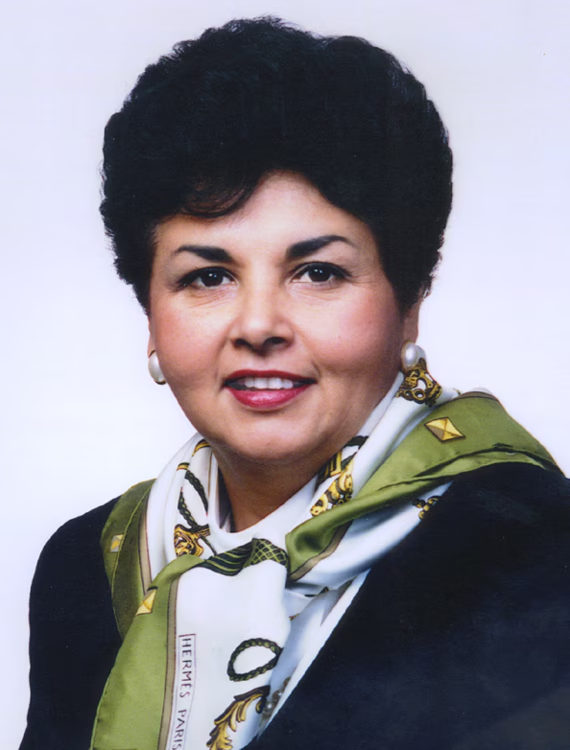 A headshot of Ana Sol Gutierrez, dressed in a black blazer with a green and white scarf.