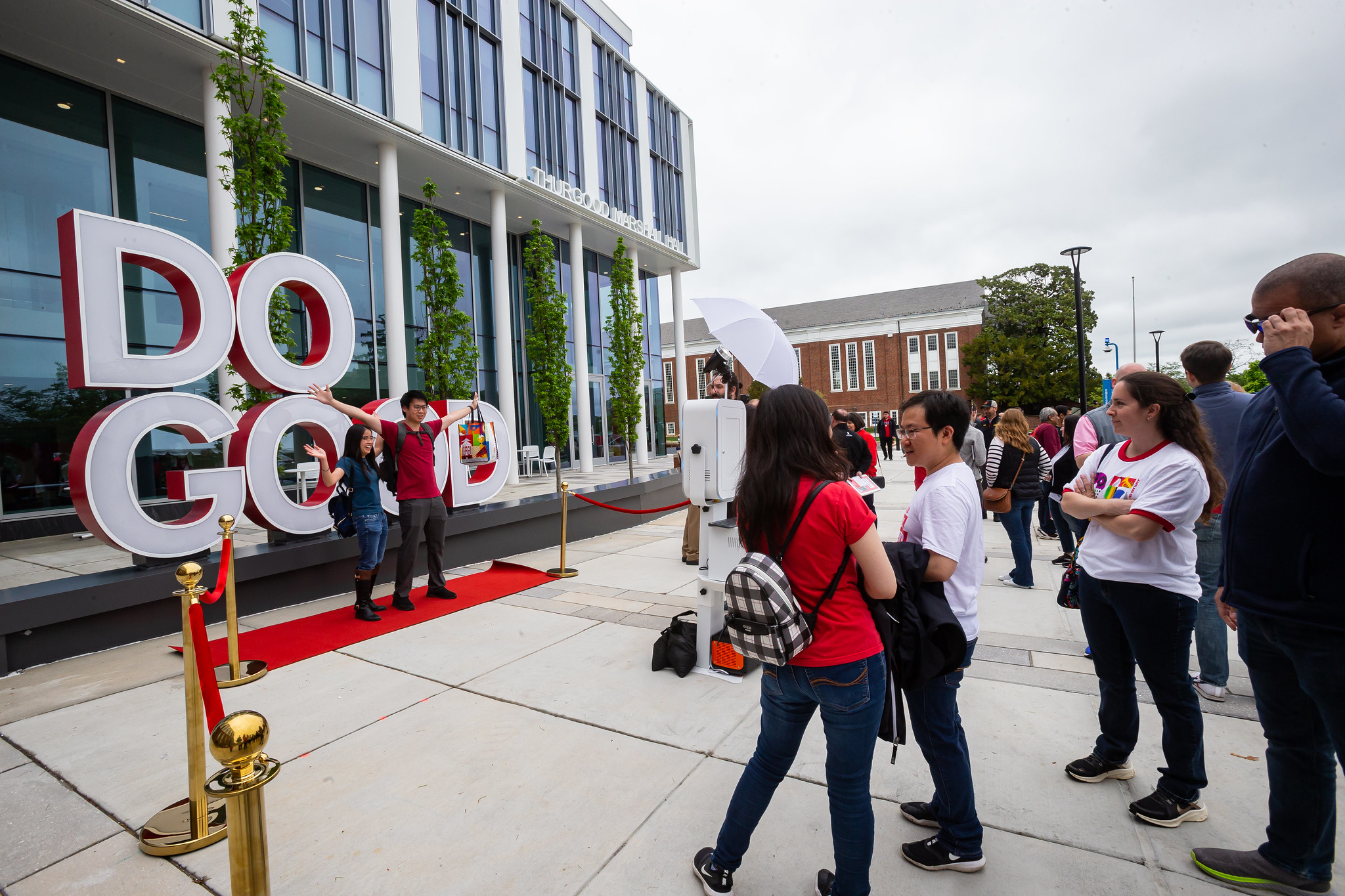 A man and a woman are standing with arms raised in front of a large Do Good sign. 