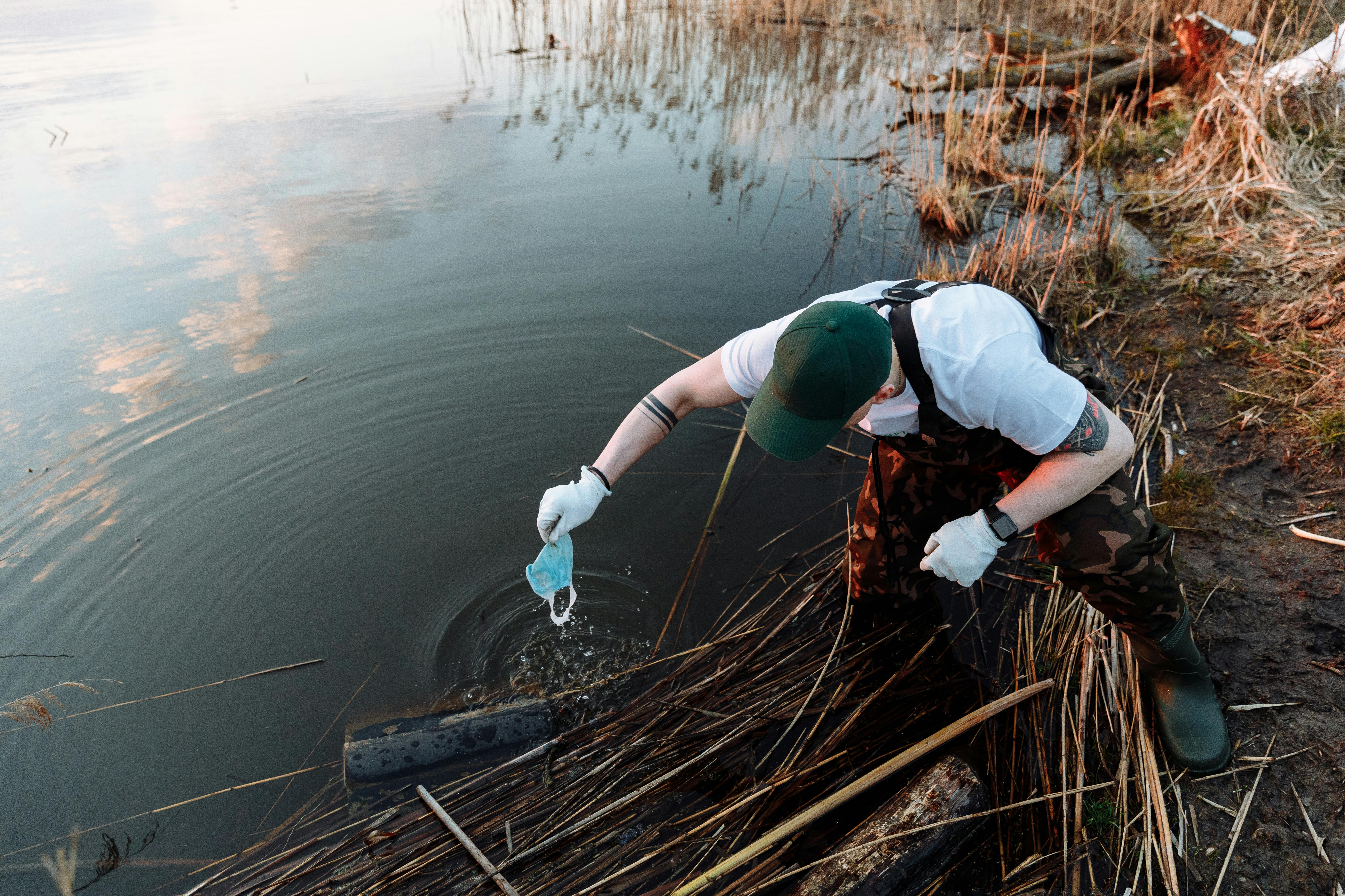 A man is leaning into a river to get some trash out of the water