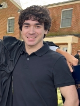 Liam Trump's headshot features him with black hair and a black shirt posed for a phot with a brick building with two windows behind him.