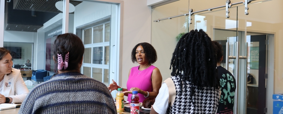A woman in a purple dress is addressing a group of students at a high top table.