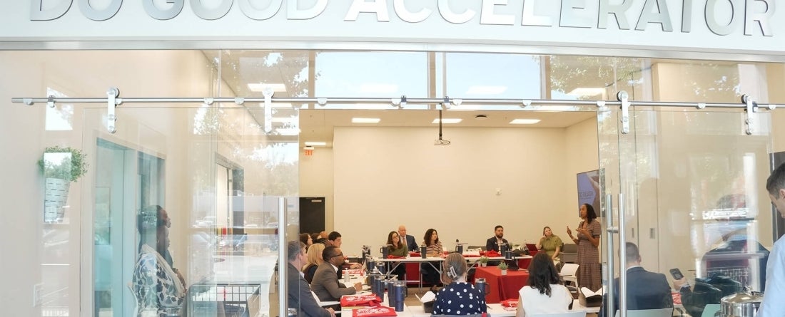 People talking behind a glass window in a well lit room. A sign above the glass window reads "Do Good Accelerator"