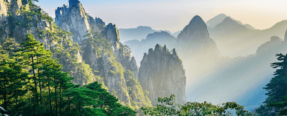 Mountains on a foggy day with blue skies and green trees in the foreground.