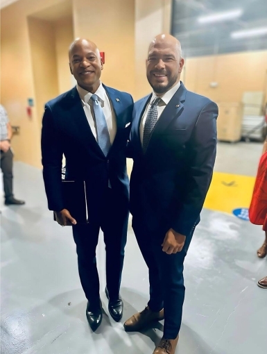 Wes Moore and John Ronquillo stand against a yellow backdrop.